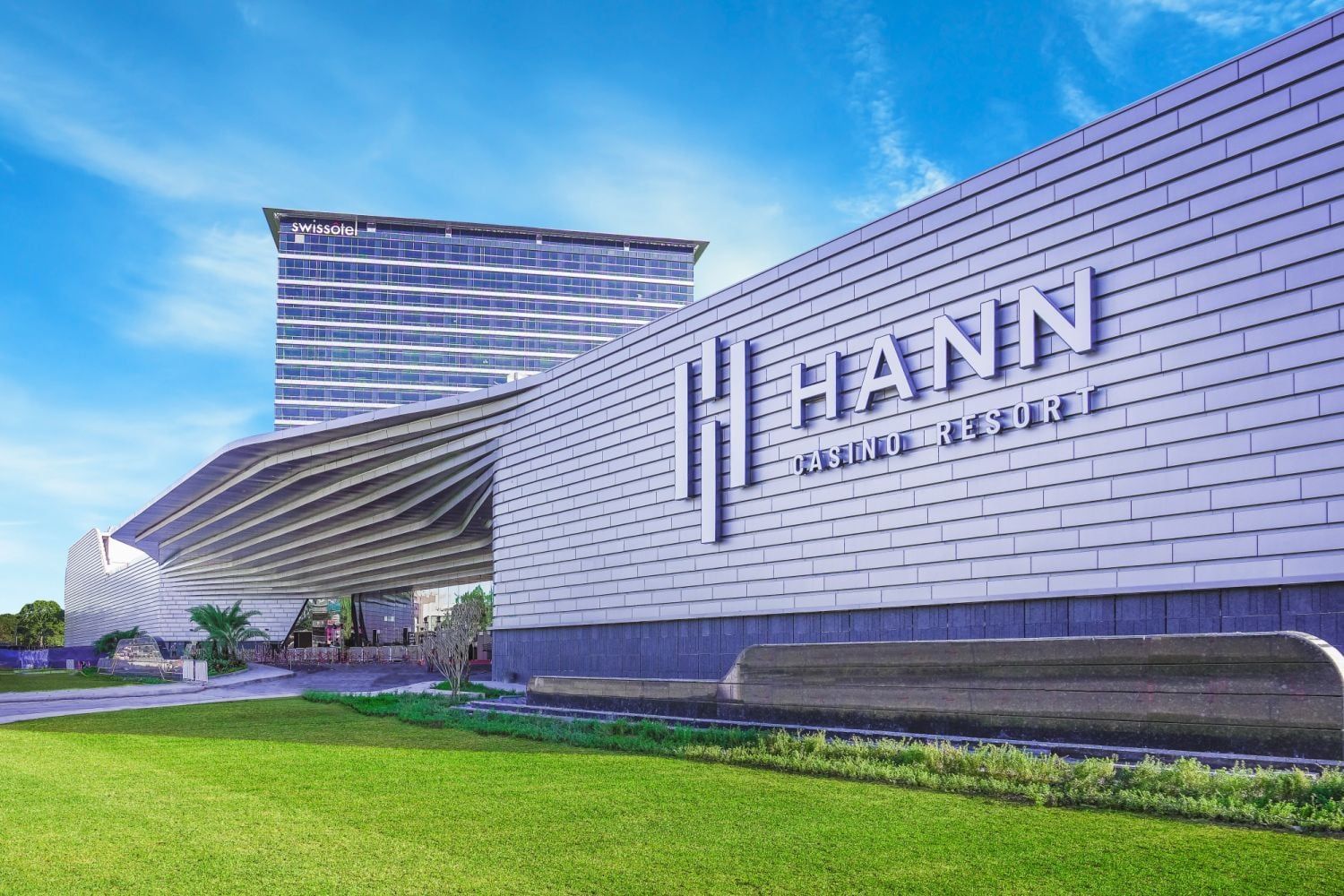 The curved, ribbed facade of Hann Casino Resort in Clark under a bright sky, its landscaped forecourt in the foreground.