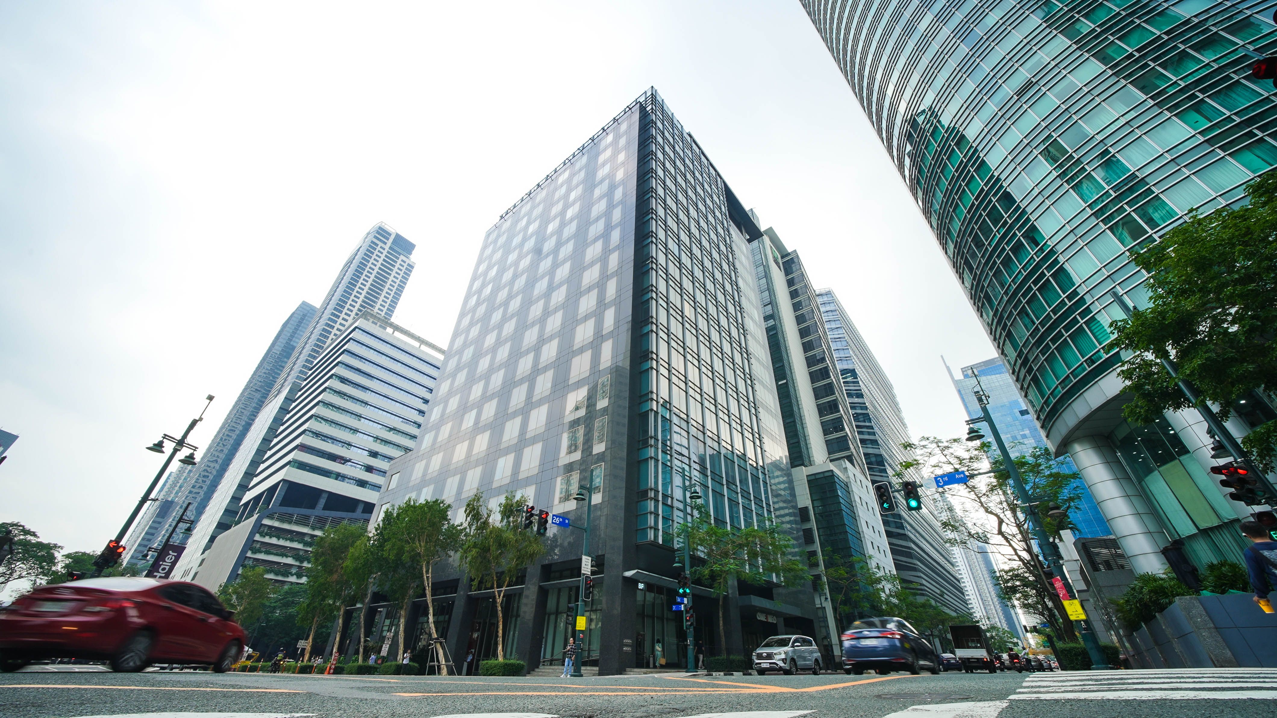 Glass-clad office towers at NEO in Bonifacio Global City, seen from street level against a bright sky.