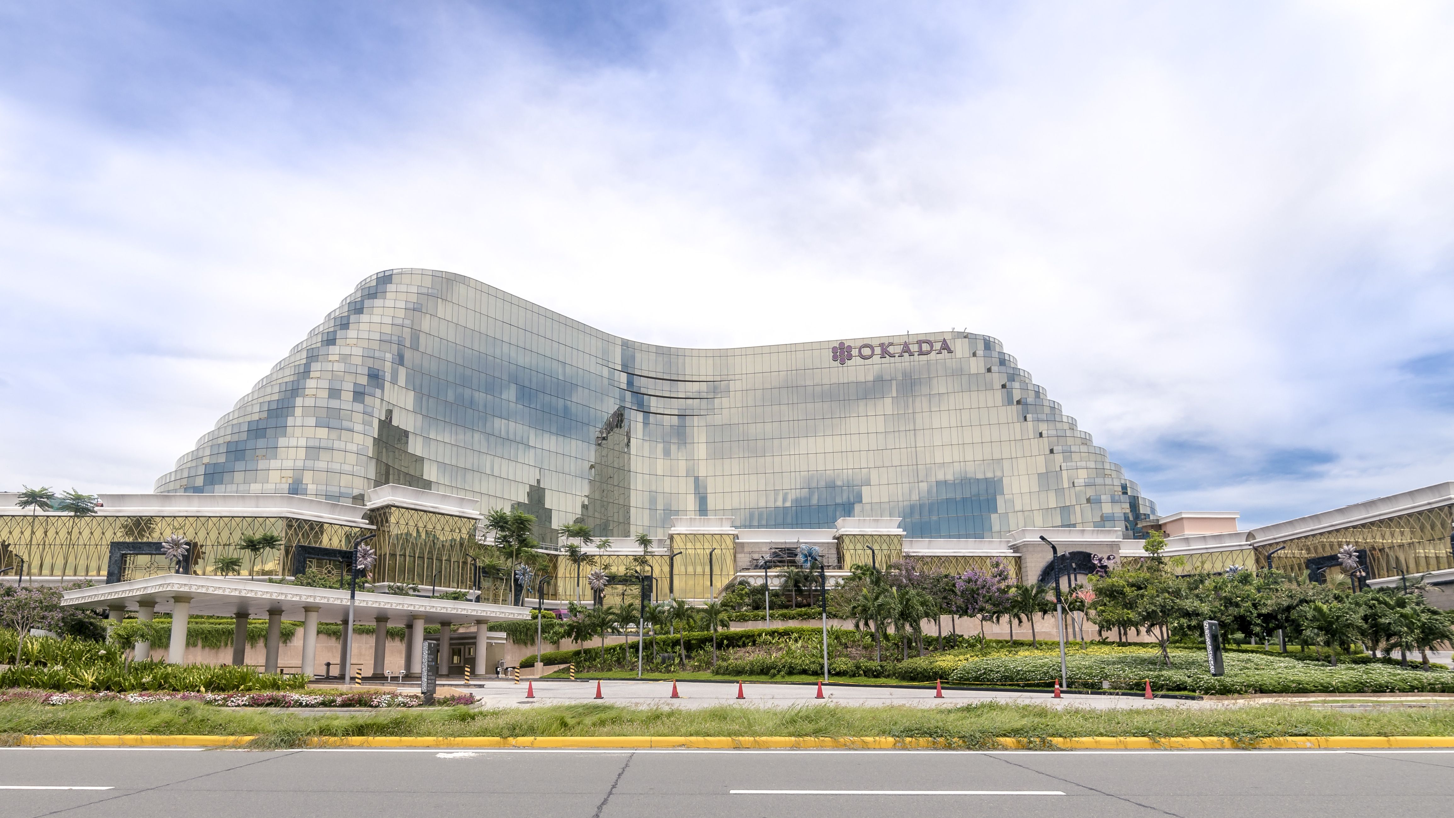 The curved glass and gold facade of Okada Manila seen from the approach road under a bright sky.